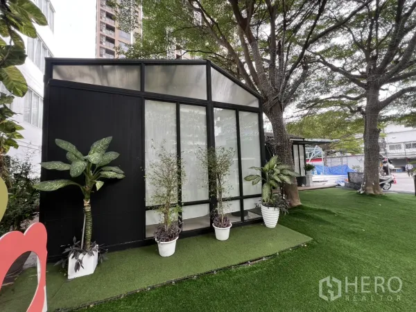 Retail Space for rent in Watthana, Bangkok - Compact glasshouse-style unit with tall windows, potted plants and shaded artificial turf beside mature trees.
