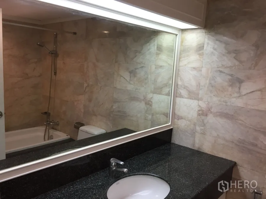Condominium for rent in Khlong Toei, Bangkok - Bathroom with marble-tiled walls, bathtub with shower, and a long granite vanity with large mirror.