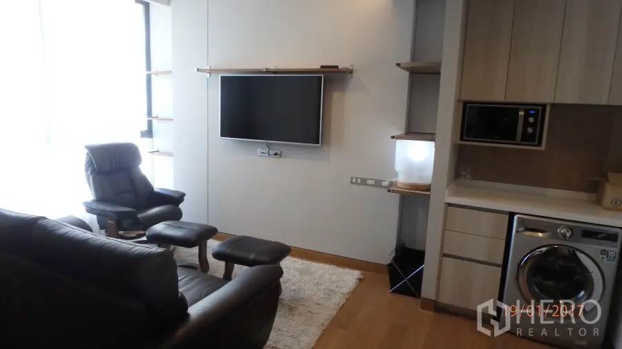 Condominium for rent in Khlong Toei, Bangkok - Modern living room of a Bangkok condo with leather lounge chair, wall‑mounted TV, large window, wood floor, and kitchenette with washing machine.