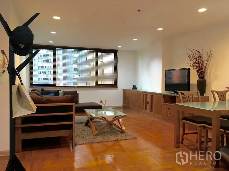 Condominium for rent in Pathum Wan, Bangkok - Bright open-plan living and dining room with parquet floors, sectional sofa, glass coffee table, and large window with city view.