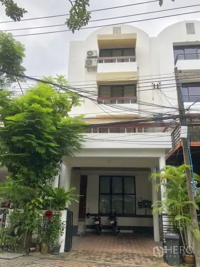 Townhouse for rent or sale in Bangkok - Four-storey white townhouse facade with covered carport, trees and a paved driveway in Watthana.