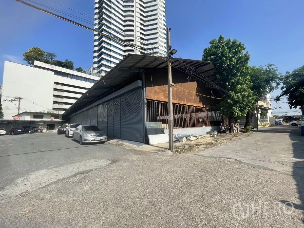 Warehouse for rent or sale in Yan Nawa, Bangkok - Street view of a single-storey warehouse with metal shutters and parking area beside high-rise buildings in Yan Nawa.