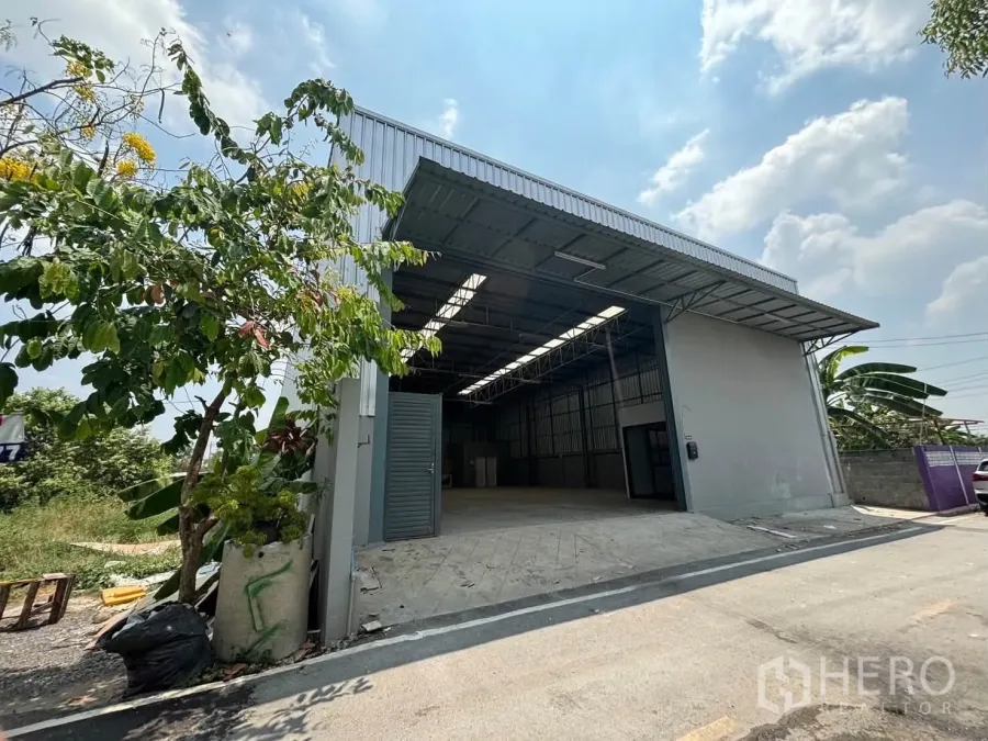 Warehouse for rent in Mueang Pathum Thani, Pathum Thani - Exterior view of the warehouse showing a wide entrance, driveway ramp, and metal roof canopy.
