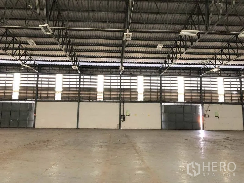 Warehouse for rent in Khlong Luang, Pathum Thani - Interior wall lined with louvered vents, steel roof trusses, and multiple entry doors.