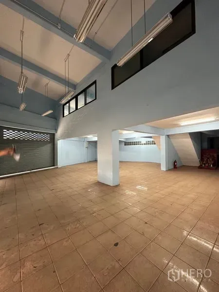 Shophouse for rent in Bang Na, Bangkok - Spacious ground-floor hall with high ceiling, columns, fluorescent lights and a roller shutter.