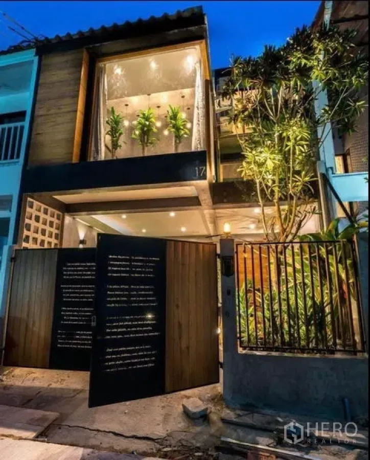 Townhouse for rent in Khlong Toei, Bangkok - Modern two-storey townhouse facade at dusk with wood gate, glass frontage and lush plants by the entrance.
