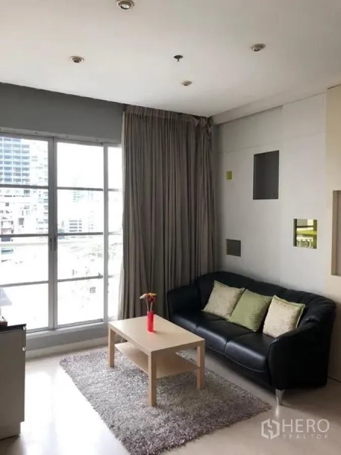 Condominium for rent in Khlong Toei, Bangkok - Bright living room with floor-to-ceiling windows, grey curtains, a black leather sofa, and a light wood coffee table on a rug with city views.