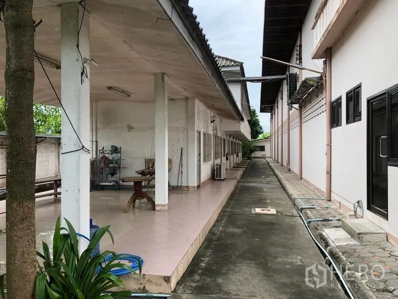 Factory for sale in Lam Luk Ka, Pathum Thani - Shaded veranda and staff facilities along a concrete access lane between buildings.