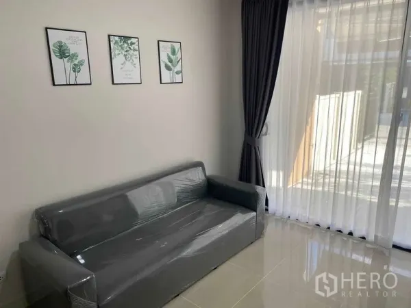 Townhouse for rent in Prawet, Bangkok - Sunlit living area with gray sofa, wall art and floor-to-ceiling curtains.