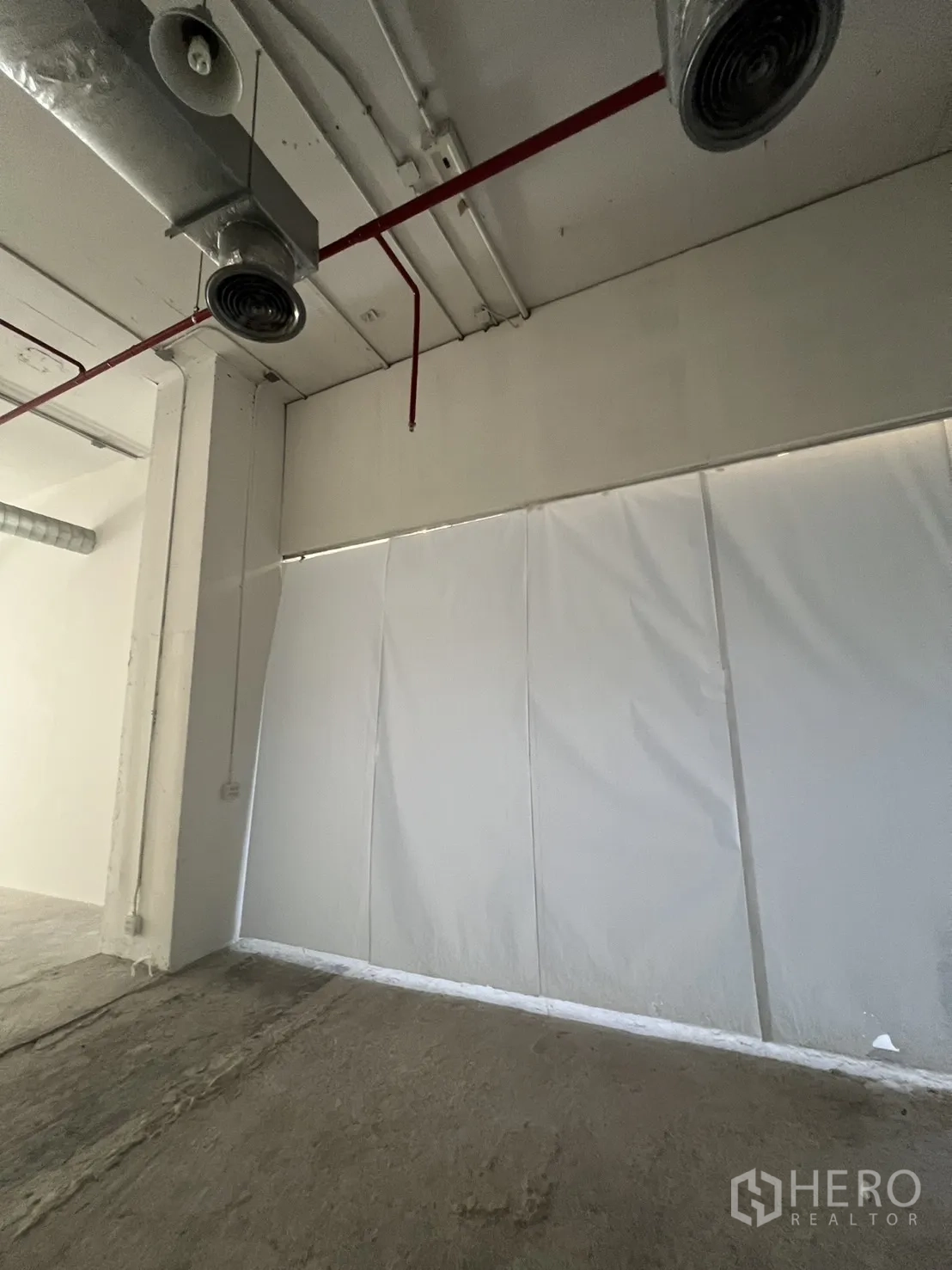 Retail Space for rent - Side wall with temporary hoarding, exposed red sprinkler pipe, ventilation outlets, and unfinished concrete floor.