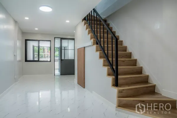 Townhouse for sale in Watthana, Bangkok - Bright living room with marble-look floor and a wooden staircase with black metal railing leading upstairs.