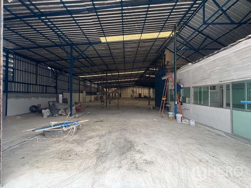 Warehouse for rent in Prawet, Bangkok - Entrance view of the warehouse showing concrete flooring, corrugated metal walls, a ladder, and a glass-partitioned office block.