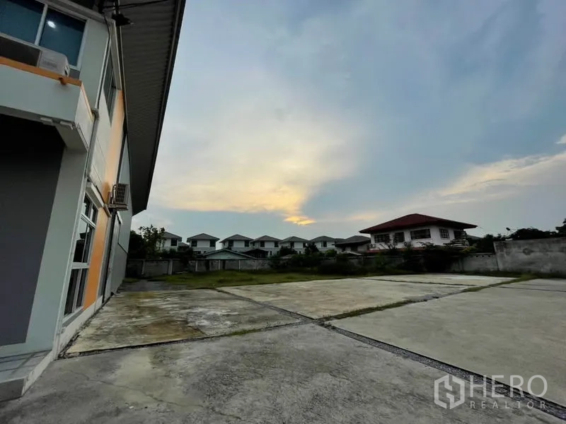 Warehouse for rent in Bang Kapi, Bangkok - Large paved yard beside the warehouse at dusk with neighboring houses in the background.