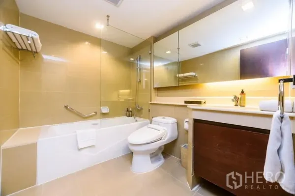 Condominium for rent in Khlong Toei, Bangkok - Bright bathroom with bathtub and glass shower screen, large mirror and vanity storage.