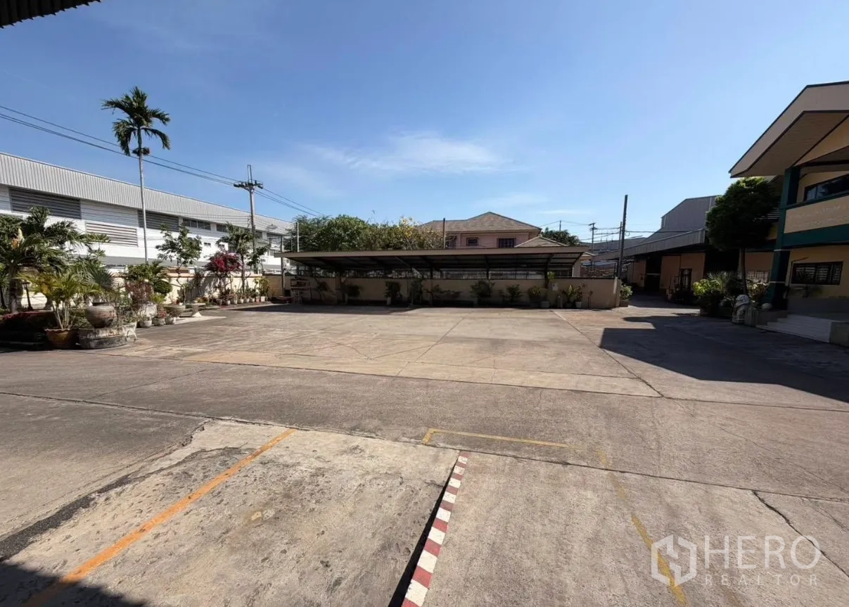 Factory for rent in Bang Phli, Samut Prakan - Wide concrete yard with covered parking and tropical plants at an industrial compound.