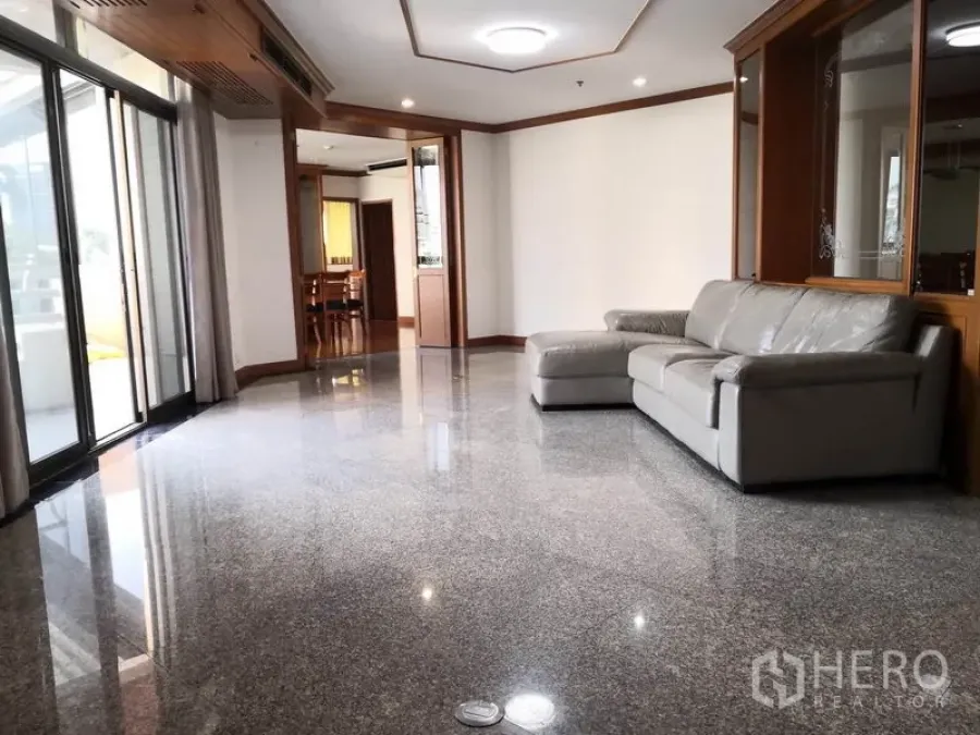 Condominium for rent in Khlong Toei, Bangkok - Bright living room with polished stone floors, L-shaped leather sofa, recessed lighting and large sliding doors to the balcony.
