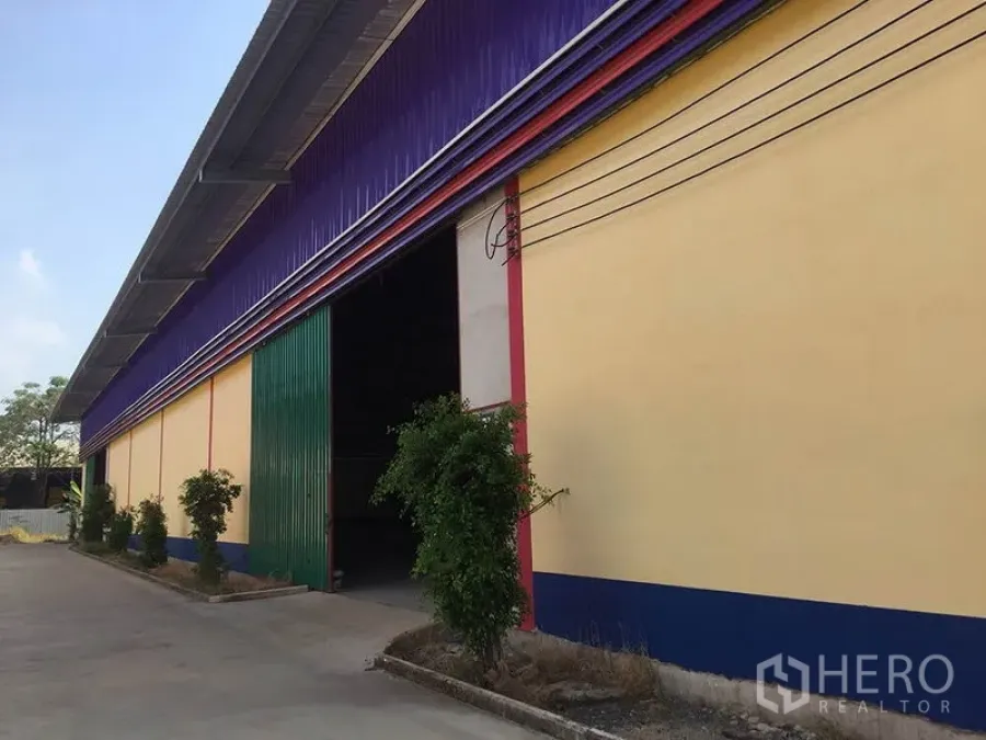 Warehouse for rent in Sai Noi, Nonthaburi - Warehouse facade with green sliding door, purple cladding and small landscaped shrubs.