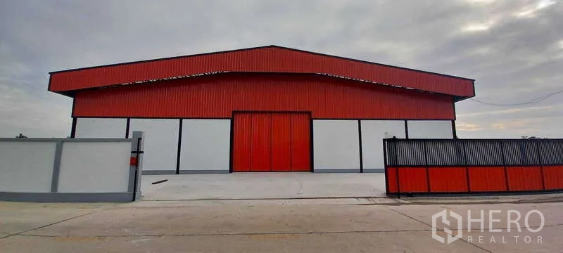 Warehouse for rent or sale in Khlong Luang, Pathum Thani - Daytime facade of a red-and-white warehouse with a gated entrance, high roof and concrete apron.
