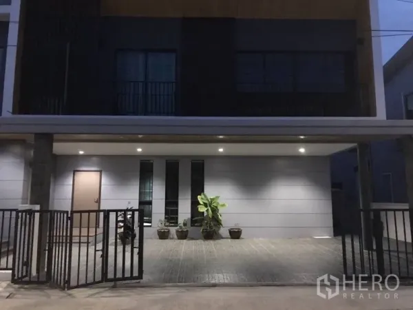 独立式住宅 出租在Thanyaburi, Pathum Thani - 黄昏时的现代独栋别墅外立面，带有有顶车位、庭院大门和盆栽。