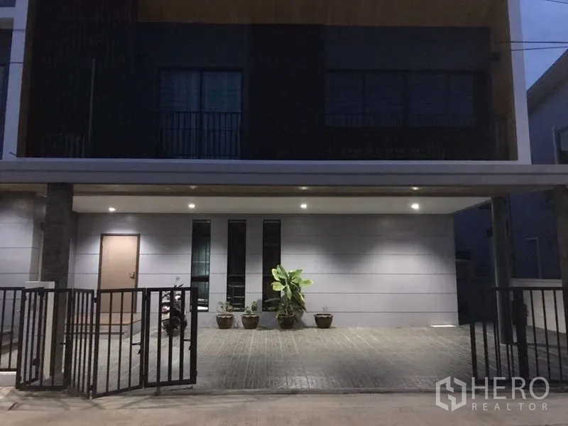 独立式住宅 出租在Thanyaburi, Pathum Thani - 黄昏时的现代独栋别墅外立面，带有有顶车位、庭院大门和盆栽。