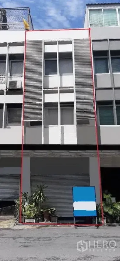 Shophouse for rent in Watthana, Bangkok - Modern multi-storey shophouse facade with a ground-floor roller shutter, vertical louver panels and potted plants, daytime street view in the Thonglor area.