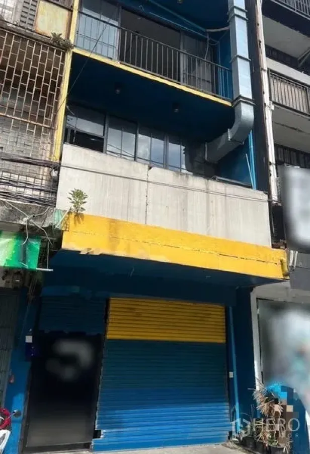 Shophouse for rent in Watthana, Bangkok - Multi-storey shophouse exterior with blue facade, yellow awning and roll-up shutter.