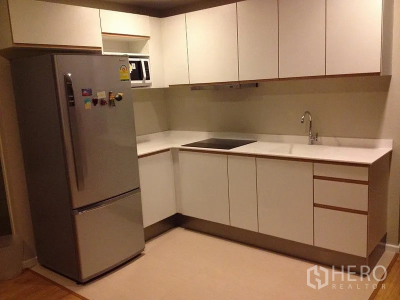 Condominium for rent in Pathum Wan, Bangkok - Modern L-shaped kitchen with white cabinets, induction hob, microwave and stainless fridge.