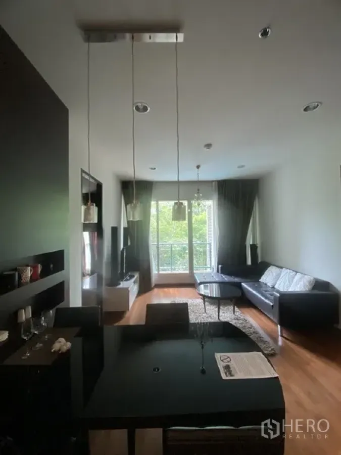 Condominium for rent in Pathum Wan, Bangkok - Open-plan living and dining room with pendant lights, black table, sofa, TV console, hardwood floors, and a balcony window facing greenery.