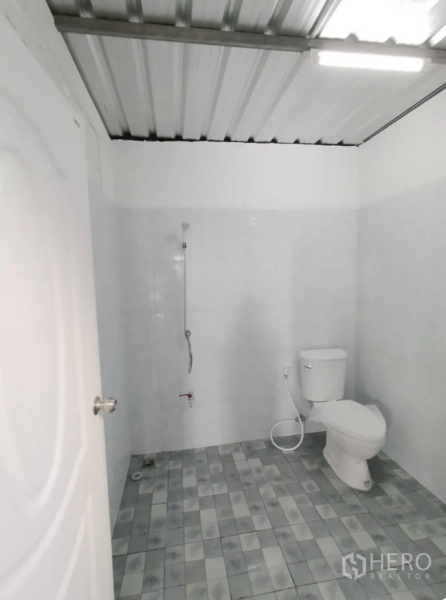 Warehouse for rent in Bang Khen, Bangkok - Clean warehouse restroom with white tiled walls, handheld shower, and ceramic toilet on a patterned tile floor.