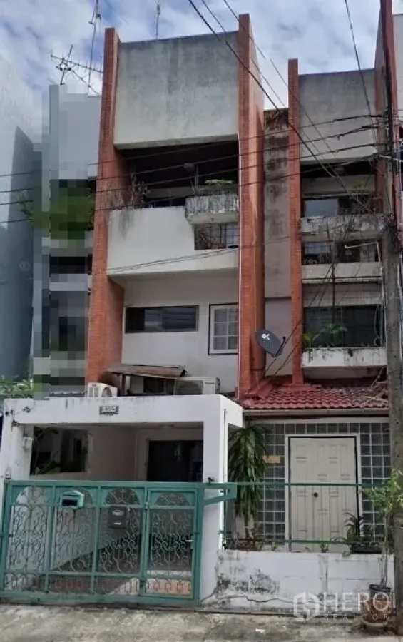 Shophouse for sale in Khlong Toei, Bangkok - Street-facing multi-storey shophouse in Bangkok with red brick columns, balconies with plants, a green metal gate, gated carport, and overhead utility lines.