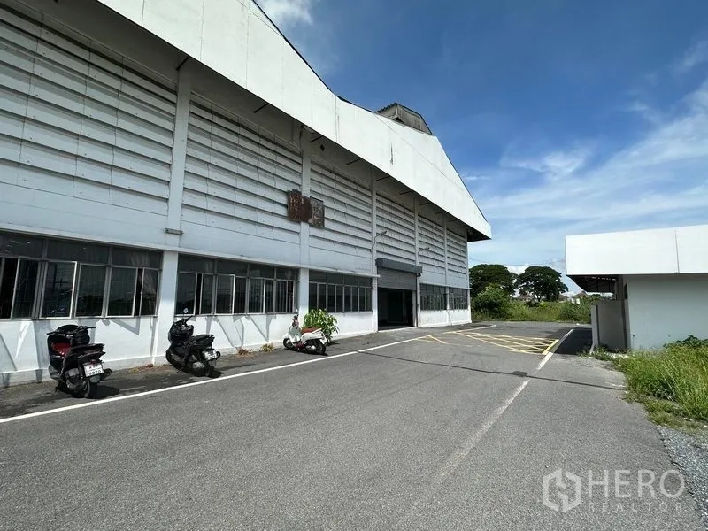 货栈 出租在Lam Luk Ka, Pathum Thani - 大型白色仓库外立面，带装卸入口与宽阔车道，墙边停有摩托车，天空晴朗。