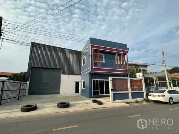 Warehouse for rent in Lam Luk Ka, Pathum Thani - Front elevation of metal-clad warehouse and modern blue office with driveway and roadside access.