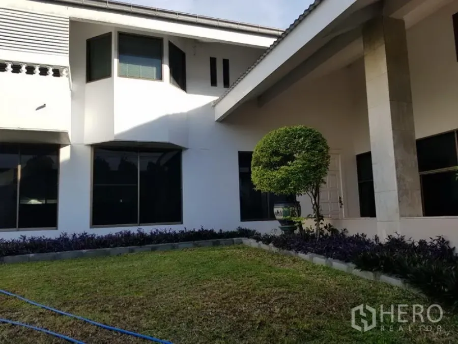 Detached House for rent or sale in Suan Luang, Bangkok - Two-storey white detached house exterior with large windows, covered porch columns, and a manicured lawn with a rounded topiary.