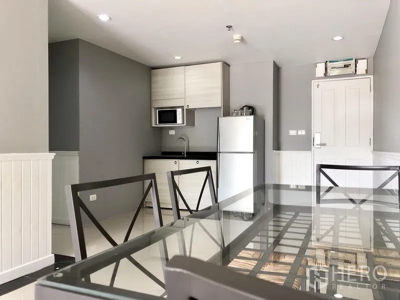 Condominium for rent in Khlong Toei, Bangkok - Compact kitchen with microwave, storage cabinets and full-size refrigerator beside a dining table.