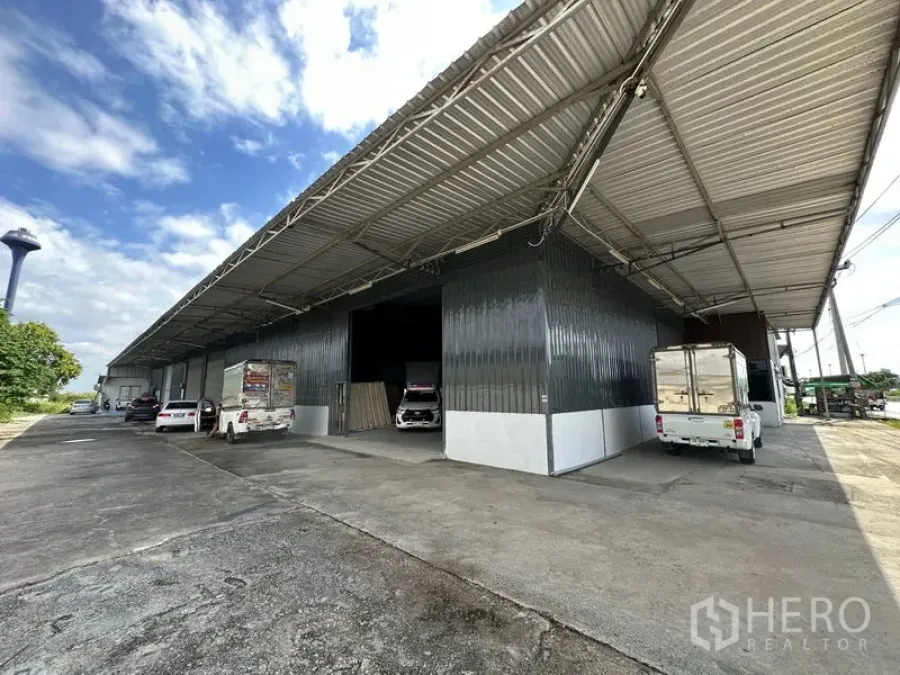 Warehouse for rent in Khlong Luang, Pathum Thani - Large metal-clad warehouse with a wide covered loading bay and vehicles parked along the driveway.