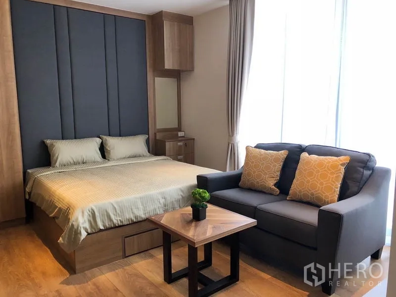 Condominium for rent in Khlong Toei, Bangkok - Bedroom with upholstered headboard, sofa and coffee table beside a floor-to-ceiling window.