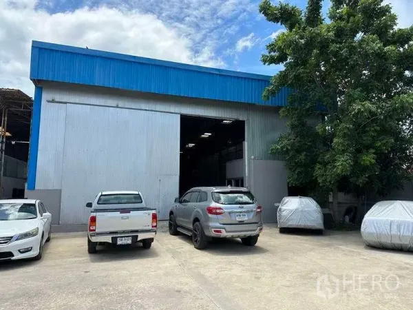 Warehouse for rent in Mueang Pathum Thani, Pathum Thani - Warehouse facade with wide roller door, on-site parking and mature tree for shade.
