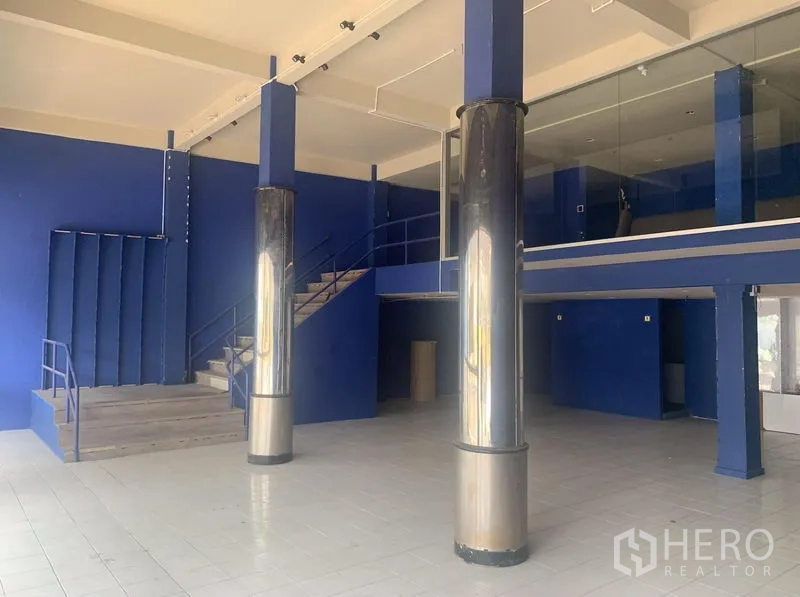 Shophouse for rent in Watthana, Bangkok - High-ceiling showroom area with metal columns, stairs, and a glass-fronted mezzanine.