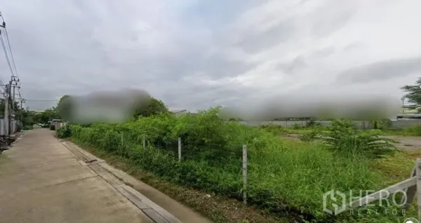 Land for rent in Prawet, Bangkok - Corner view of fenced vacant land next to a paved road with utility lines and neighboring buildings in Prawet, Bangkok.