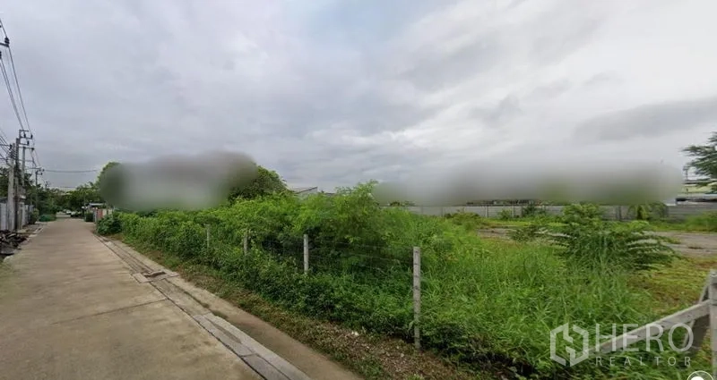 Land for rent in Prawet, Bangkok - Corner view of fenced vacant land next to a paved road with utility lines and neighboring buildings in Prawet, Bangkok.