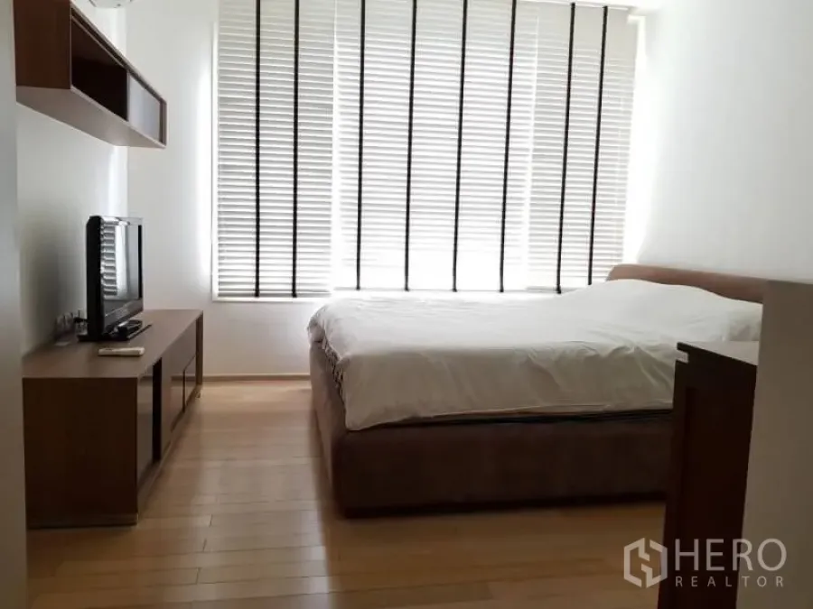 Condominium for rent in Khlong Toei, Bangkok - Bright bedroom with large window blinds, wood floors, queen bed, and TV console.