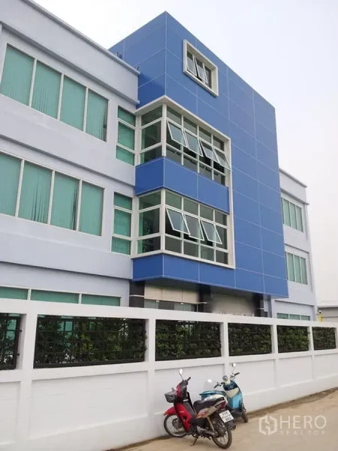 Warehouse for rent or sale in Bang Phli, Samut Prakan - Modern blue-and-white four-storey warehouse and office facade with corner glazing behind a white boundary wall.