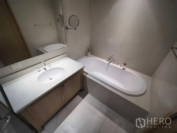 Condominium for rent in Khlong Toei, Bangkok - Contemporary bathroom with white vanity, large mirror and built-in bathtub.