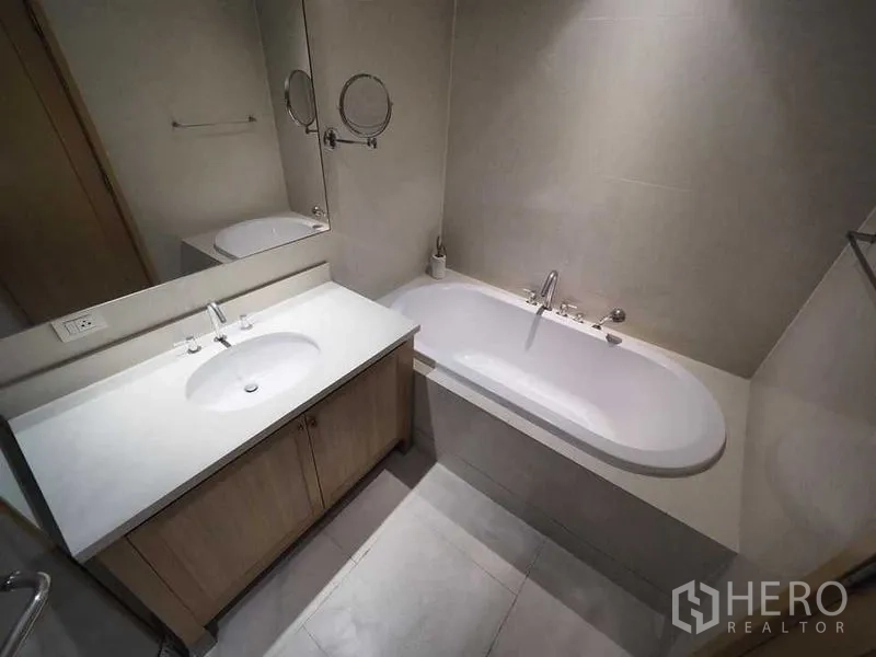 Condominium for rent in Khlong Toei, Bangkok - Contemporary bathroom with white vanity, large mirror and built-in bathtub.