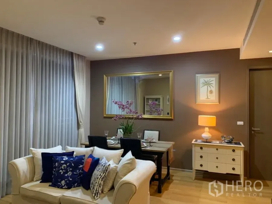 Condominium for rent in Watthana, Bangkok - Warm living and dining area with cream sofa, set dining table, large wall mirror and soft ambient lighting.