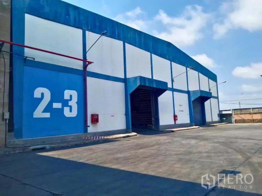Factory for rent in Samut Prakan - Blue and white factory exterior with roller shutter loading bays and wide truck driveway.