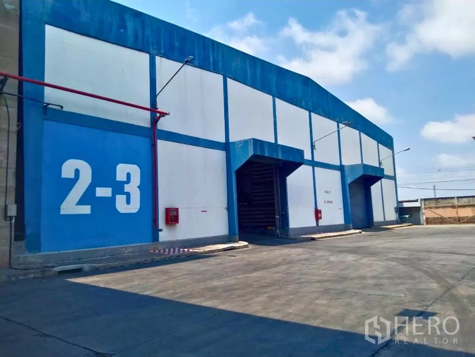 Factory for rent in Samut Prakan - Blue and white factory exterior with roller shutter loading bays and wide truck driveway.