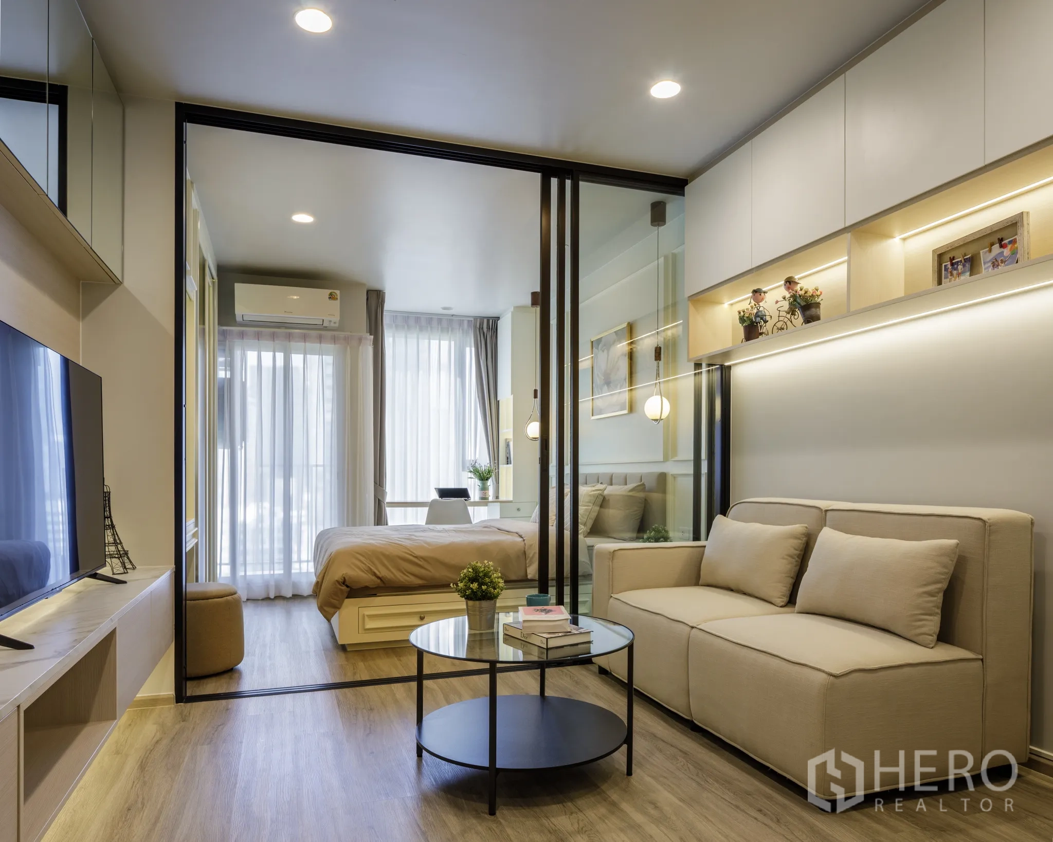 Condominium for rent in Khlong Toei, Bangkok - Bright living room with beige sofa, TV console, round coffee table, and sliding glass doors opening to a bedroom with desk and floor-to-ceiling windows.