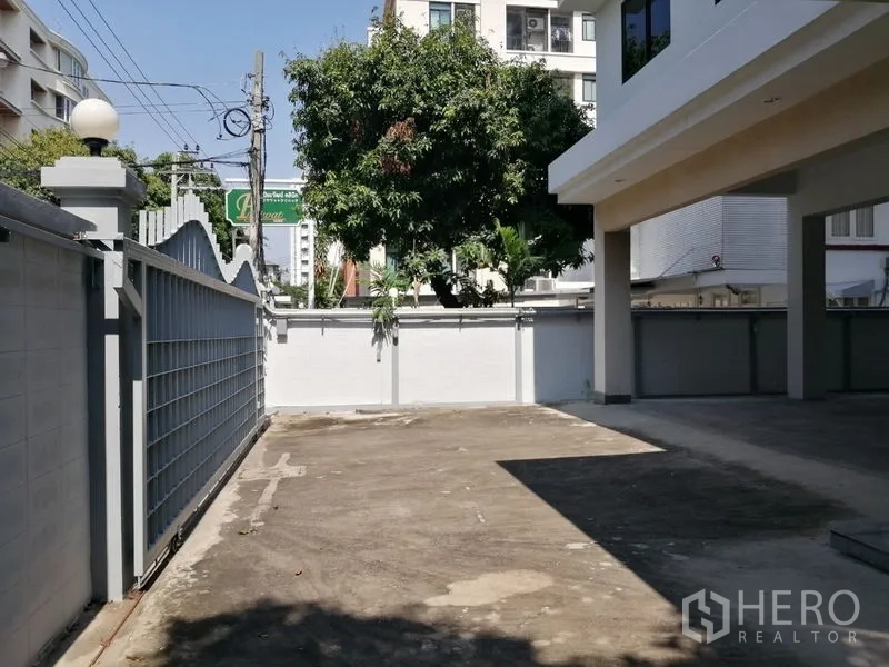 Home Office for rent in Watthana, Bangkok - Gated driveway and open parking area beside the building with a mature tree.