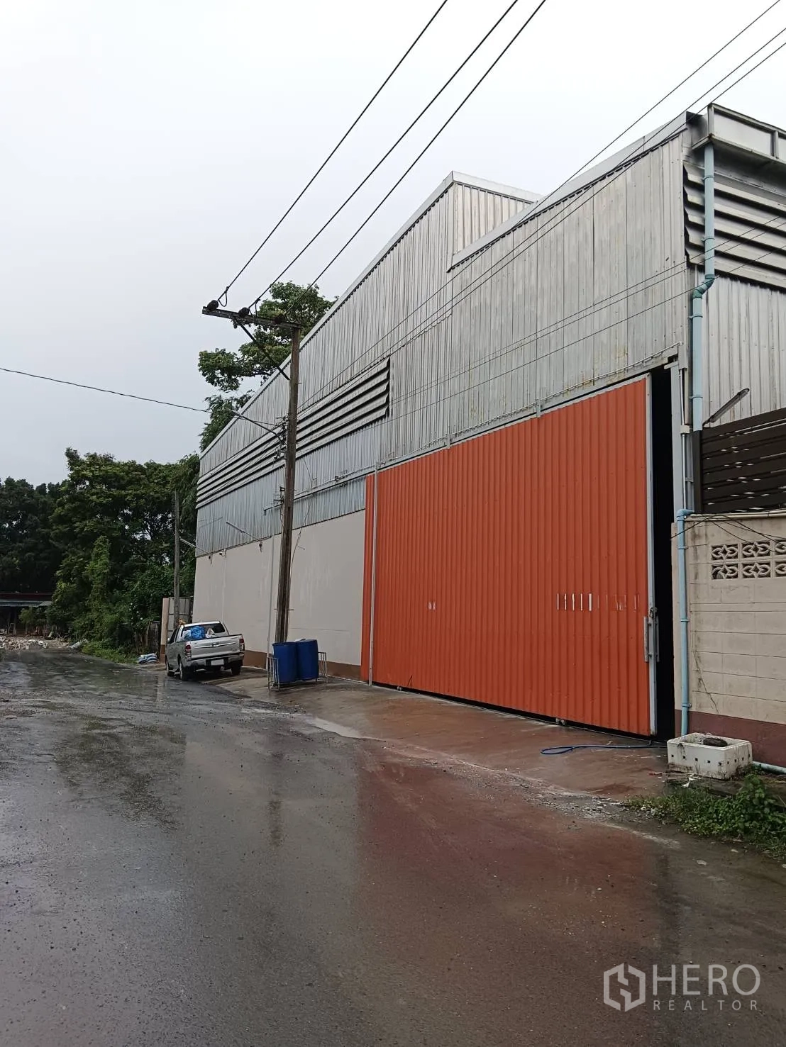 货栈 出租在Khlong Sam Wa, Bangkok - 曼谷Khlong Sam Wa仓库外立面，金属板墙体与橙色大滑门，雨后街景。